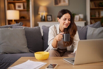 A young woman sits on a sofa, sipping coffee from a mug while using a laptop. A yellow bowl sits nearby, and soft lighting creates a warm atmosphere in the room.