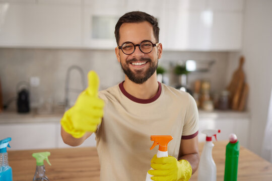 A man is smiling and giving a thumbs up while holding a spray bottle. He is wearing yellow gloves in a clean, bright kitchen filled with cleaning supplies. - Powered by Adobe