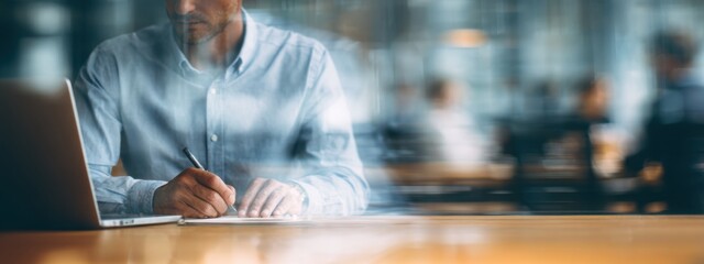 a businessman in a shirt is working on a laptop computer he taking notes or participating in a meeting in a conference room, using the laptop for a financial concept. the background is blurred.