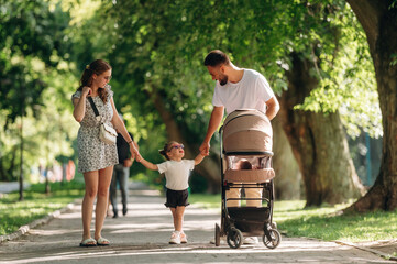 Holding girl by hands. Couple is with daughter and cradle are having a walk