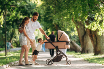 Fototapeta premium Holding girl by hands. Couple is with daughter and cradle are having a walk