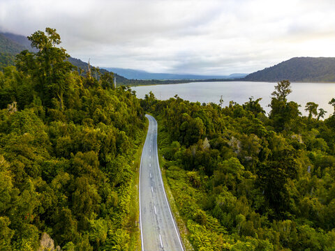 Aerial view of a winding road cutting through dense green forest leading to a serene lake under a soft, cloudy sky, Lake Brunner, West Coast, New Zealand.