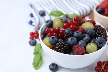 Different ripe juicy berries and basil in bowl on white tiled table, closeup