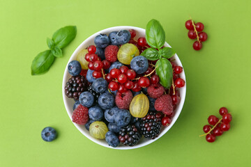 Different ripe juicy berries and basil in bowl on green background, flat lay