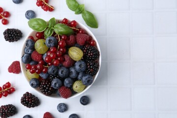 Different ripe juicy berries and basil in bowl on white tiled table, flat lay. Space for text