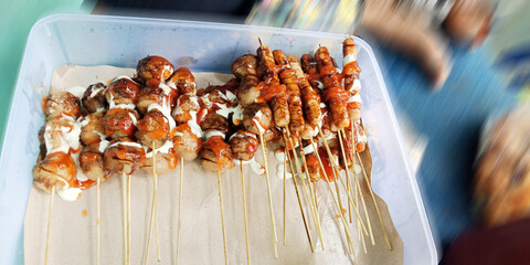 An overhead view of meatball skewers and sausage skewers in a plastic container, drizzled with spicy red sauce and creamy white sauce.