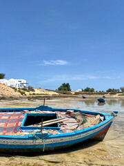Obraz premium Fishing boats rest along the quiet shoreline of Djerba in Tunisia. The blue sky reflects on calm waters, providing a peaceful scene of seaside life and fishing activity.