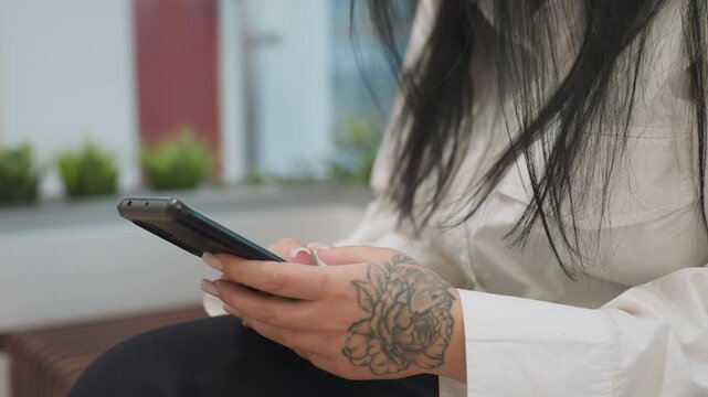 Partial view of woman with floral tattoo on hand using smartphone while seated, surrounded by blurred decorative green plants in background, dressed in white shirt and black pants