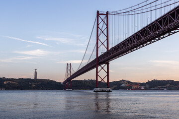 View of the vibrant red 25 de Abril Bridge stretches across the tranquil blue waters, connecting distant shores under a serene sky, Lisbon, Portugal.
