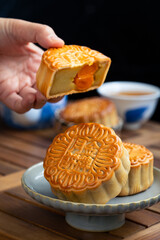Traditional Chinese moon cake in a plate with yolk inside, hand holding it about to eat with cup and tea on wooden table in black background. Chinese character on it represent "Gold medal mooncake"