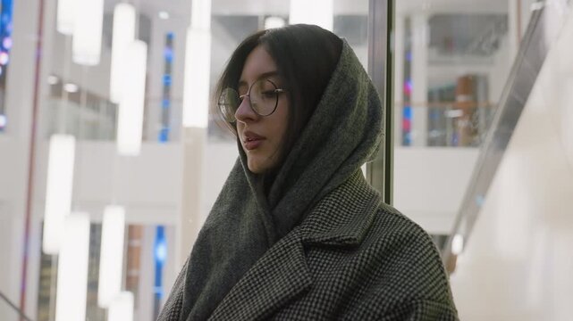 young woman in glasses and houndstooth coat stands in glass elevator sipping juice, surrounded by bright hanging lights and modern decor as elevator ascends through stylish shopping mall interior