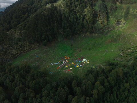 Aerial view of colourful tents dot a lush green meadow nestled amidst the dark, dense forests, creating a vibrant contrast against the mountainous backdrop, Sosan, Himachal Pradesh, India.