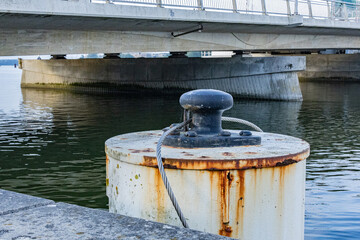 Fototapeta premium Close up of rusty mooring bollard with steel cable near concrete bridge over water