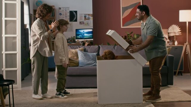 Mother covering sons eyes with her hands when they entering room where father opening lid of huge box with mini poodle inside, surprised boy and woman petting it, dog jumping out