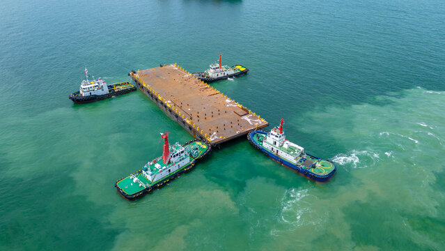 Aerial view of tugboats towing a large rusty steel barge in the ocean, used for transporting heavy industrial cargo or offshore construction. Maritime logistics and shipping concept.