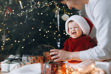 Cute baby in santa hat crying at mother with present boxes with garland lights under modern decorated christmas tree. Sad overwhelmed baby. Holiday time