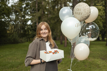 Smiling pregnant woman standing outdoors with festive balloons and a box of cupcakes during baby shower celebration. Concept of maternity lifestyle, family happiness, and pregnancy event. Symbol of