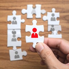 A hand holds a white jigsaw puzzle piece with a red person icon, surrounded by gray puzzle pieces with person icons on a light brown wooden surface
