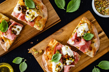 Slices of Pinsa Romana with mortadella, burrata cheese and pistachio on wooden cutting boards on black background