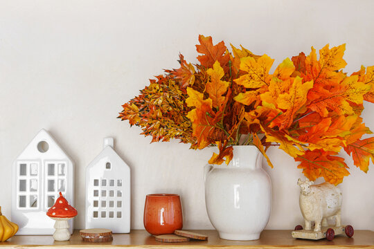 Bright orange and yellow autumn leaves in white vase on wooden table top. Nearby decorative houses and candle on white plaster wall background. Fall decor for Halloween, Thanksgiving day holidays.