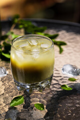 A refreshing, two-tone iced matcha beverage with ice cubes and fresh green leaves scattered on a textured table surface. This vibrant, cool drink is perfect for a warm summer day.