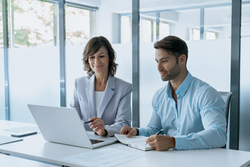 Obraz premium Team of two businesswoman and businessman working together using digital laptop pc, talking about business project. Latin mature professional ceo woman and European young student man at work in office