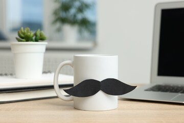 Happy Father's Day. Cup with paper moustache and laptop on wooden table, closeup