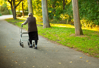 Obraz premium Elderly woman slowly taking a walk though park with help of a walker to aid 