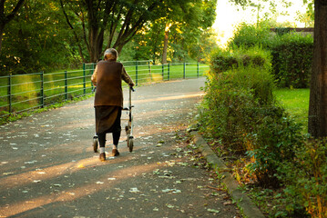 Elderly woman slowly taking a walk though park with help of a walker to aid 