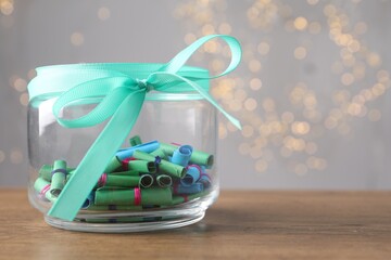 Colorful paper notes in glass jar on wooden table against grey background with blurred lights, space for text. Bokeh effect