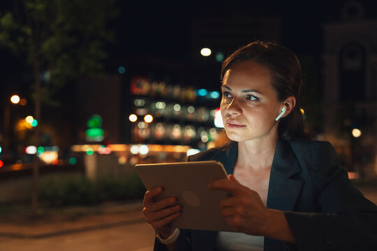 Female using digital tablet with earbuds outdoors in urban evening - Powered by Adobe