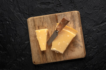 Parmesan cheese wedge on a wooden cutting board, with a cheese knife, on a black slate background, overhead flat lay shot