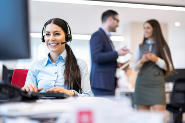 Female call center agent is working at computer wearing headset while business people are in background in work atmosphere