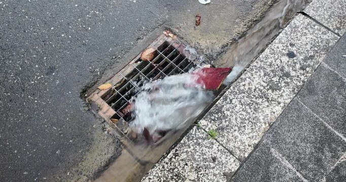 Leaves and other debris are swept by a powerful stormwater current rushing down a street gutter and into a metal storm drain.