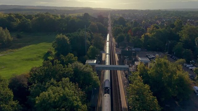 Aerial above the train departing from train station in Kent, UK. Following train journey towards Ashford on a sunny morning. Peaceful landscape with passing meadows and village. Camera facing the sun.