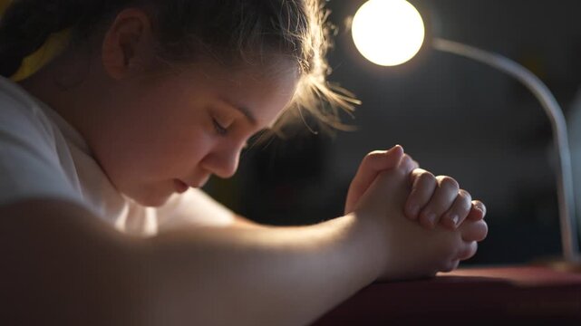 Young girl sitting at table with her hands on her head. Religious girl lifestyle praying to a child. Religious book of prayer with hands on a table. A young girl is at a table with her hands on her.
