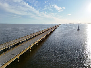 Aerial view of a straight Wright Memorial bridge crossing a tranquil sea between Kill Devil Hills and Point Harbor toward the distant horizon. The sun glints off the water, creating a serene scene.