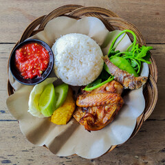 Top view of nasi tempong, a traditional Indonesian meal with rice, fried chicken, fried fish, vegetables, tempeh, and sambal served on woven bamboo plate.