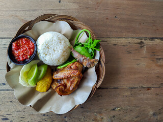 Top view of nasi tempong, a traditional Indonesian meal with rice, fried chicken, fried fish, vegetables, tempeh, and sambal served on woven bamboo plate.