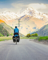 Male person in blue jacket cycling with touring bicycle surrounded by mountains and green summer...