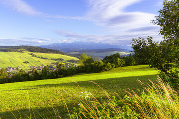 Obraz premium Sunlit hills with vibrant fields and scattered trees open toward distant blue mountains. A picturesque village nestles in the valley, embodying rural charm, traditional farming, and scenic tranquility