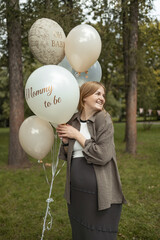 Smiling pregnant woman standing in park holding festive balloons with inscriptions Mommy to be and Oh Baby during baby shower celebration. Concept of maternity lifestyle, pregnancy happiness, and