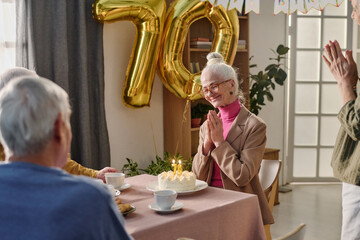 Senior Caucasian woman smiling and clasping hands while sitting at table with birthday cake and candles, celebrating seventieth birthday with friends in decorated room