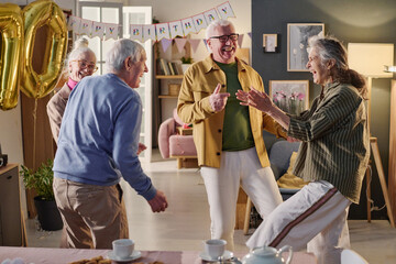 Group of senior Caucasian men and women laughing and dancing together during birthday celebration at home, large gold balloon and birthday banner visible in background, joyful interaction