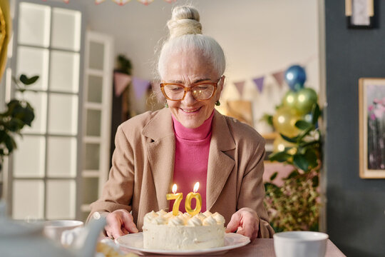 Senior Caucasian woman smiling while holding birthday cake with number seventy candles, celebrating milestone at decorated indoor party, wearing glasses and hair in bun