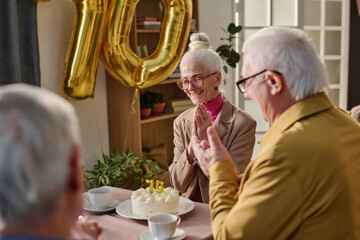 Senior Caucasian woman smiling and clasping hands while sitting at table with senior Caucasian men, celebrating birthday with cake and large gold number balloons in background