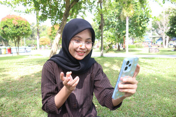 A young woman wearing a black hijab and glasses sits on a grassy lawn in a park, smiling and gesturing with her hand as she engages in a video call on her smartphone.