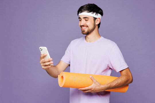 Young fitness trainer instructor sporty man sportsman wear headband t-shirt training in home gym hold yoga mat use mobile cell phone isolated on plain purple background Workout sport fit abs concept - Powered by Adobe