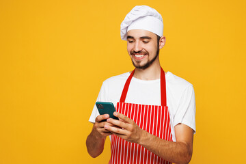 Young housewife housekeeper chef cook baker man wears red apron toque hat hold use mobile cell phone look for recipe isolated on plain yellow orange background studio portrait. Cooking food concept.