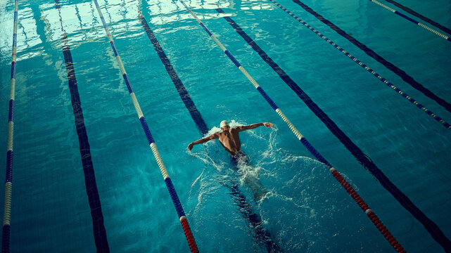 Focused swimmer executing butterfly stroke in indoor lane with powerful energy. Concept of fitness and sports advertising, motivational visuals, education on swimming technique, and wellness promotion